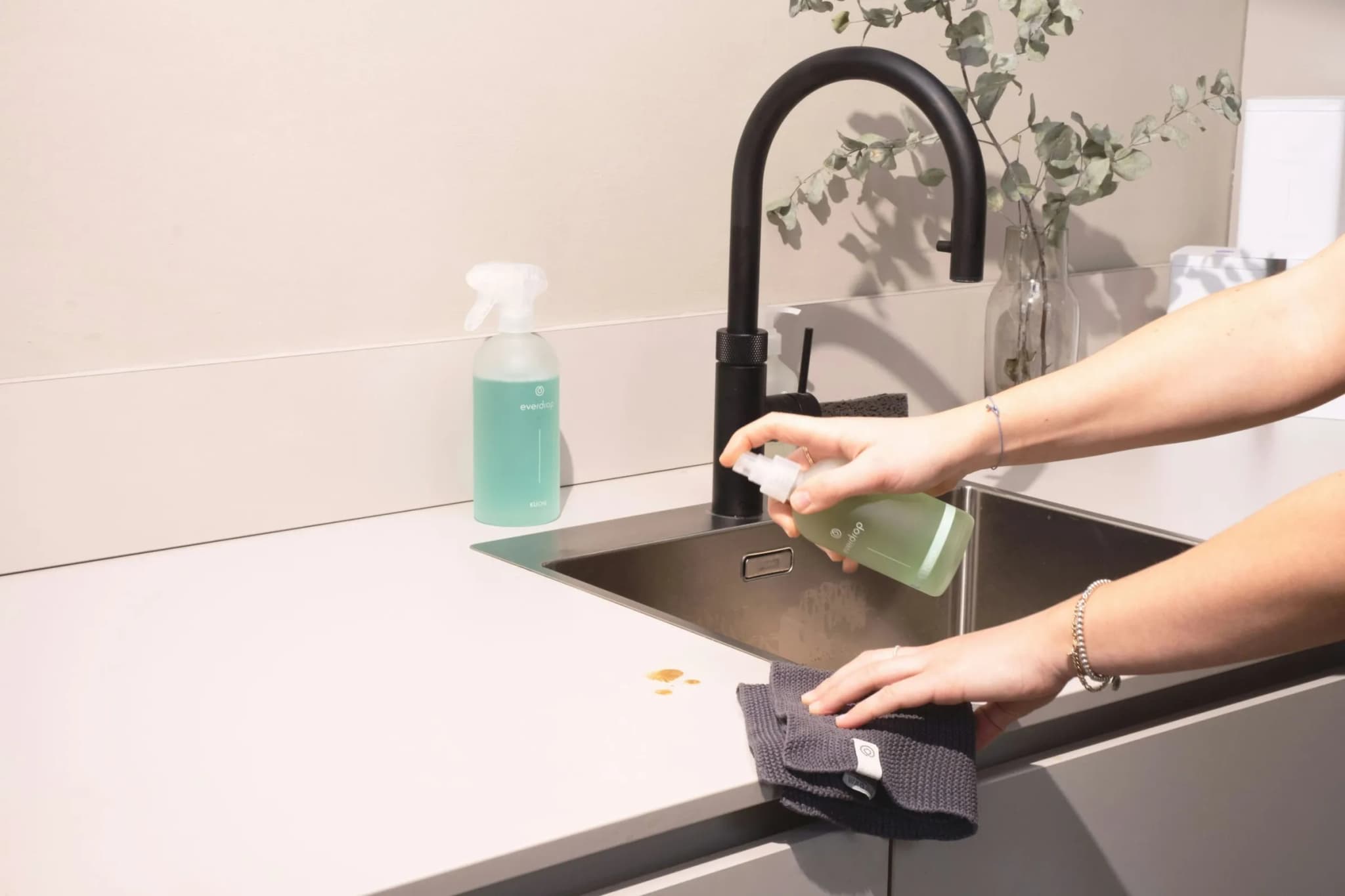 Hands cleaning a kitchen sink and worktop with spray and a cloth