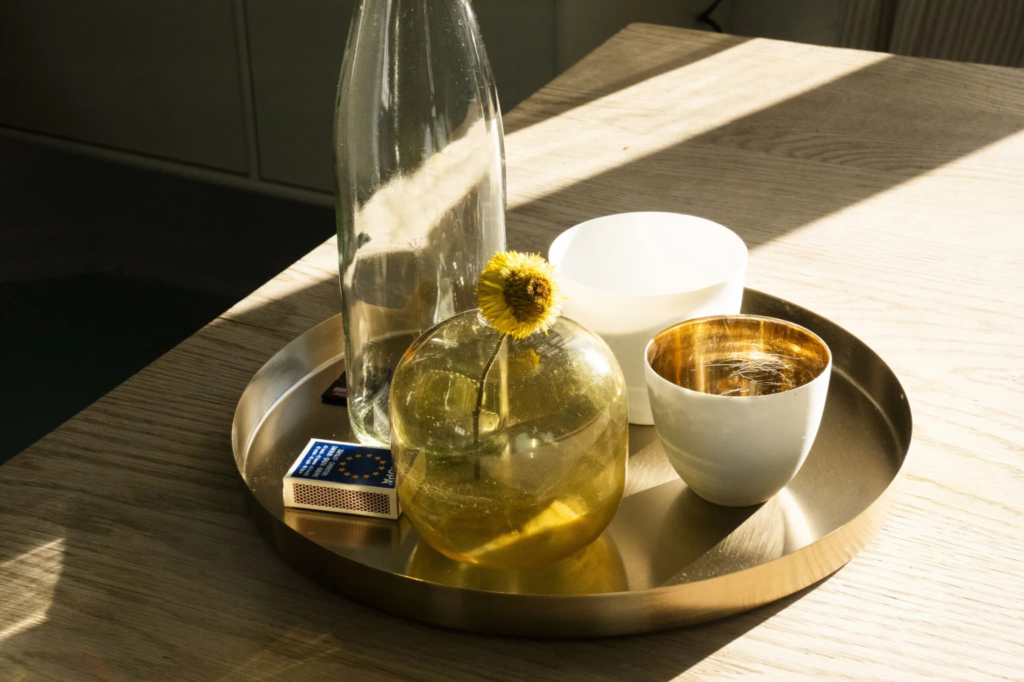 Brass tray with a glass carafe, a vase with a yellow flower and cups in sunlight