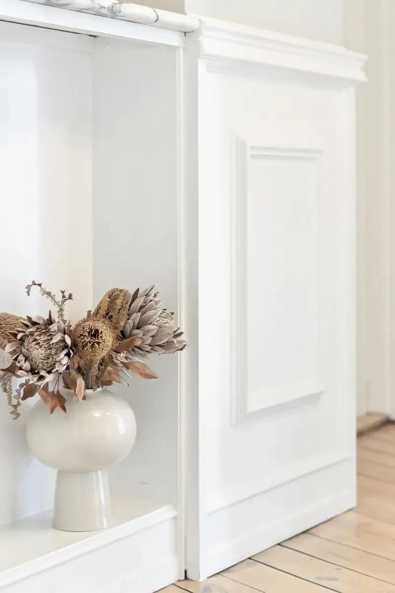 A bright room with white panelled doors, a wooden floor and a vase of dried flowers