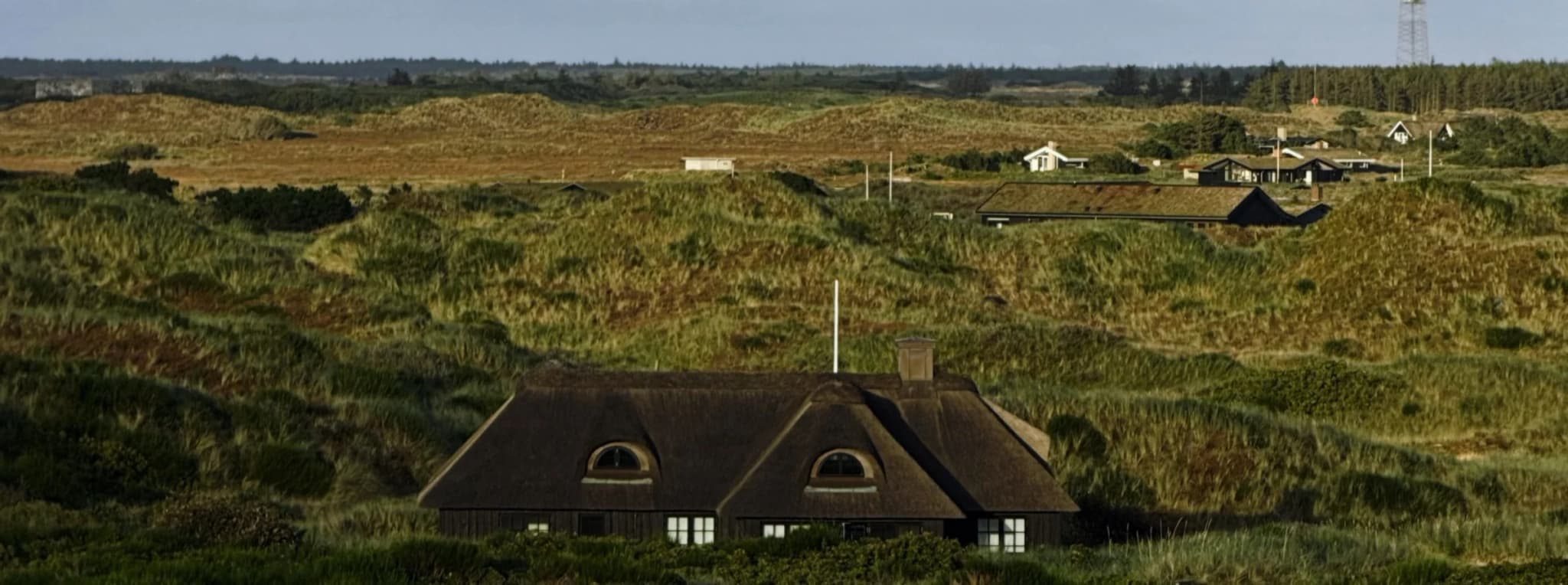 Stråtækt sommerhus i klitlandskab med et sommerhusområde og en mast i baggrunden