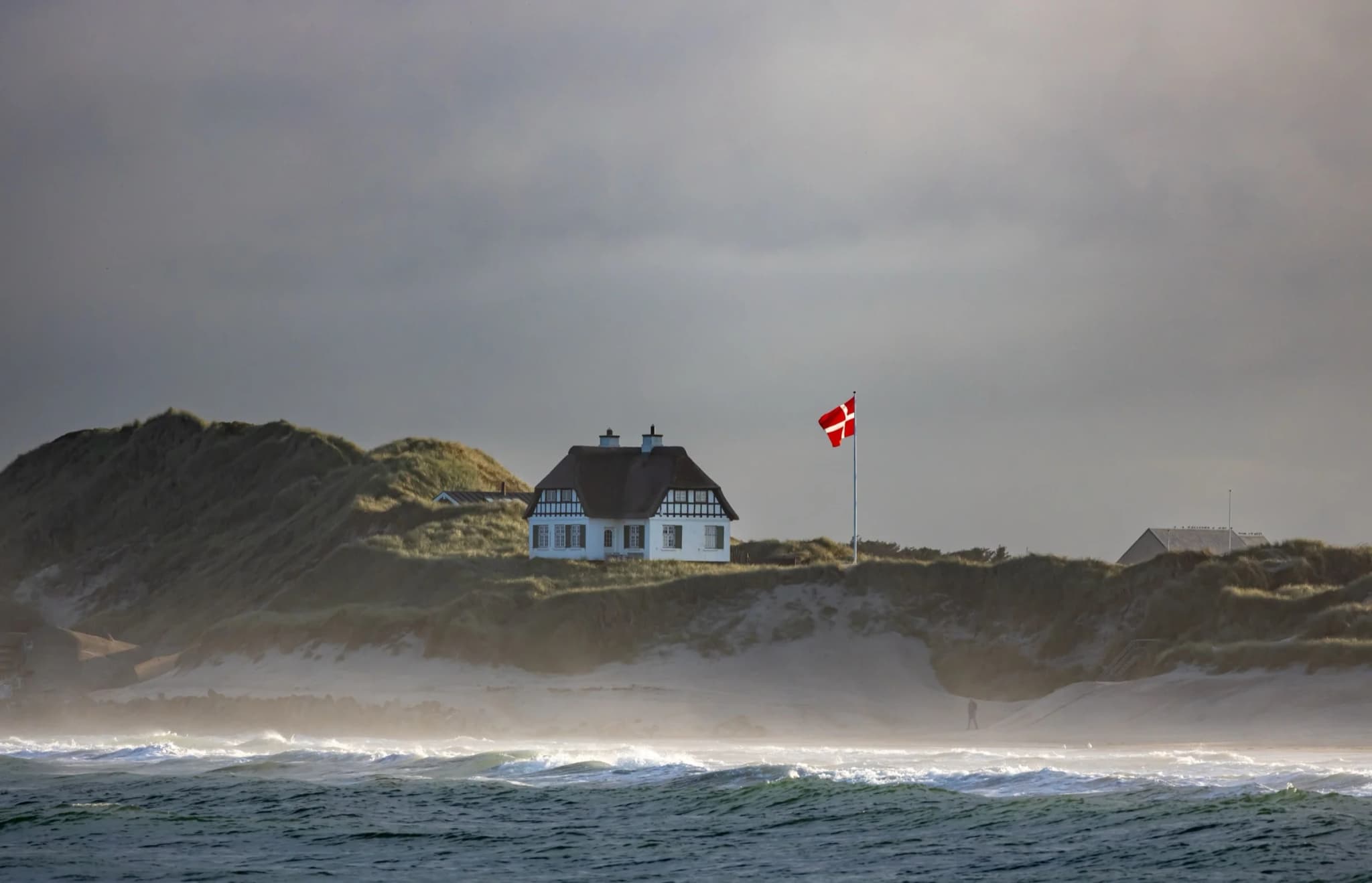 Hvidt stråtækt sommerhus på en klit ved den danske kyst med Dannebrog og bølger forrest