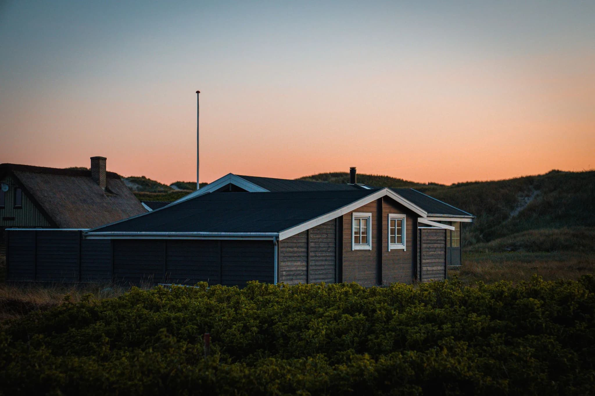 Mørkt træbeklædt sommerhus mellem klitter ved solnedgang, med et stråtækt hus i baggrunden