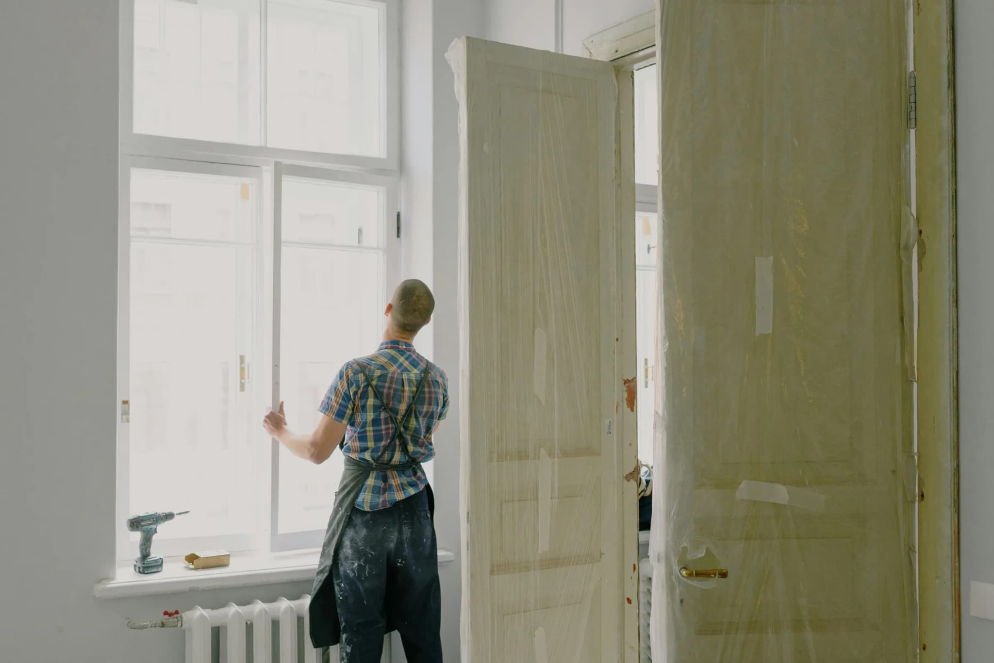 Workman in an apron, seen from behind, fitting a window next to a drill and doors in an apartment