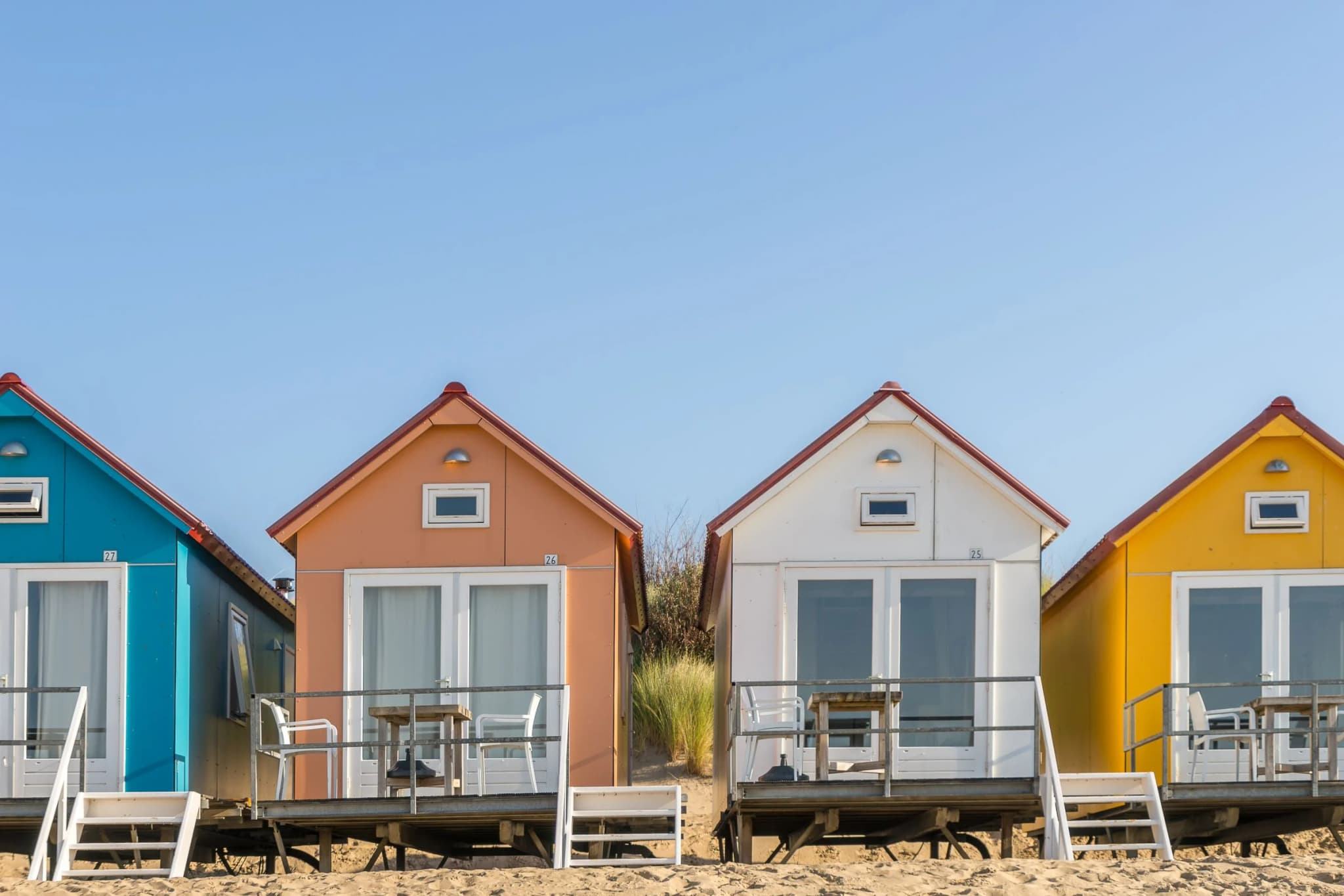 Række af ens, nummererede feriehuse i forskellige farver på en strand, side om side som i et feriecenter