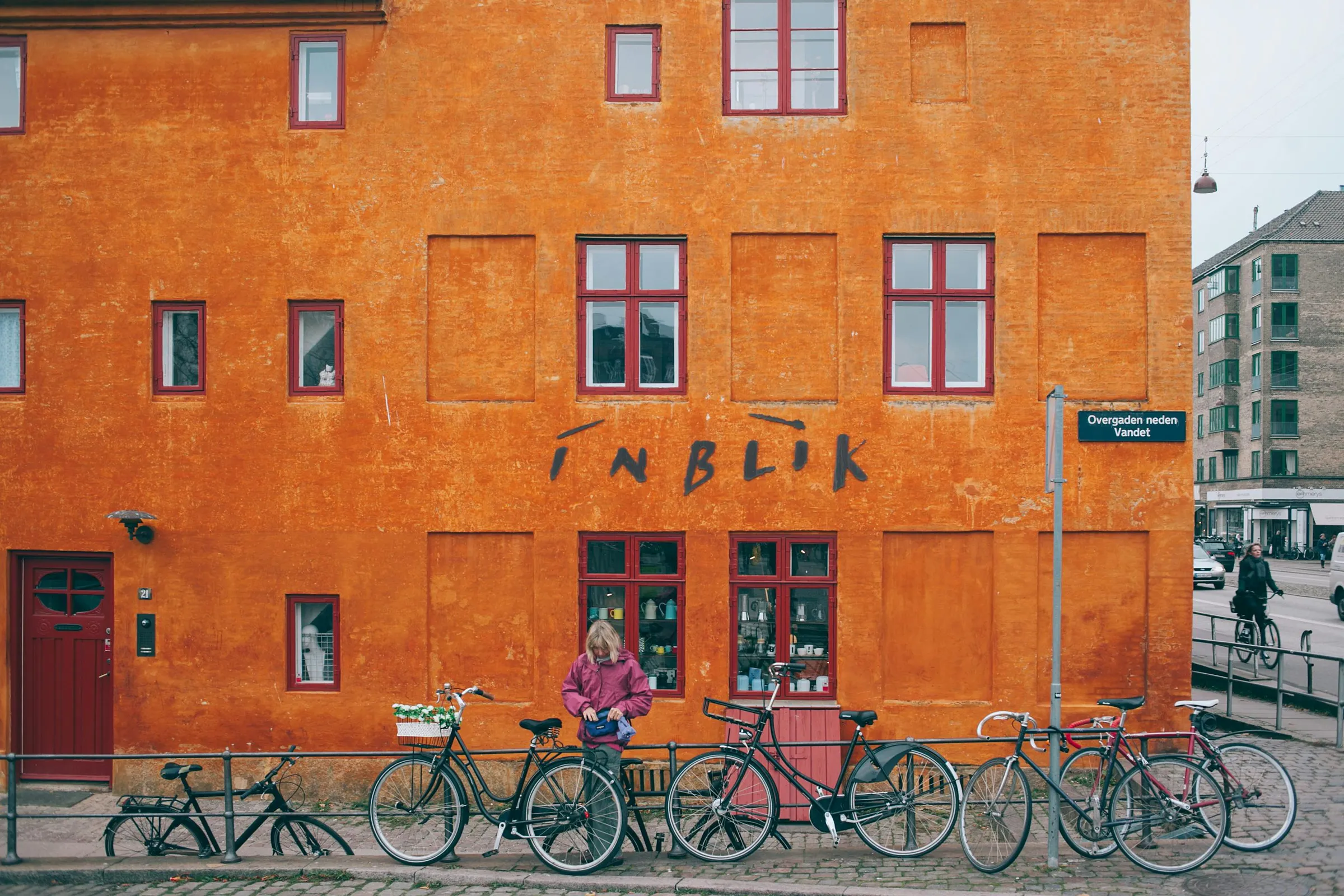 Woman parking bicycle near metal railing against colorful orange stone house with shop downstairs located in Copenhagen Denmark on cloudy day