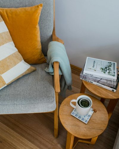 From above of comfortable armchair with colorful pillows placed near wooden side tables with books and cup of tea in living room